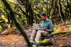 Adult man work with computer laptop and roaming phone connection in the middle of the green wild forest - concept of digital nomad and modern smart working people everywhere internet connected